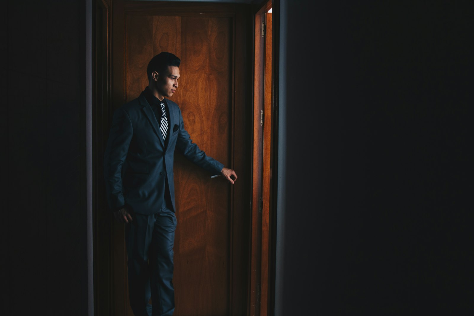 Man wearing black suit jacket and dress pants standing beside brown wooden door